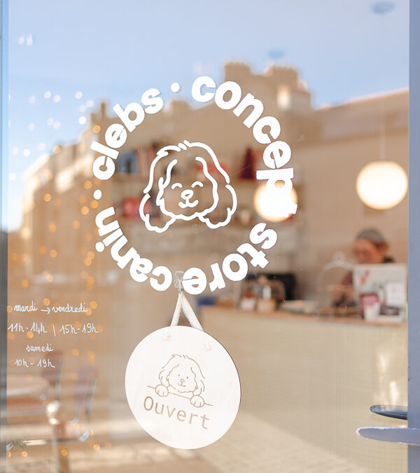 Vitre d’entrée de la boutique canine Clebs à Rennes avec le logo illustré d’un chien et une pancarte “Ouvert”, vue sur l’intérieur lumineux du concept store