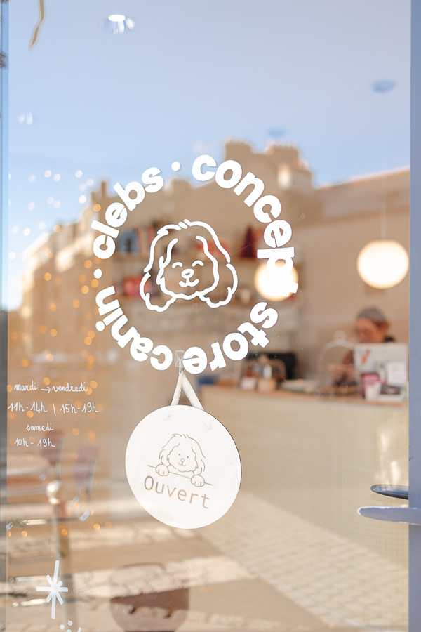Vitre d’entrée de la boutique canine Clebs à Rennes avec le logo illustré d’un chien et une pancarte “Ouvert”, vue sur l’intérieur lumineux du concept store