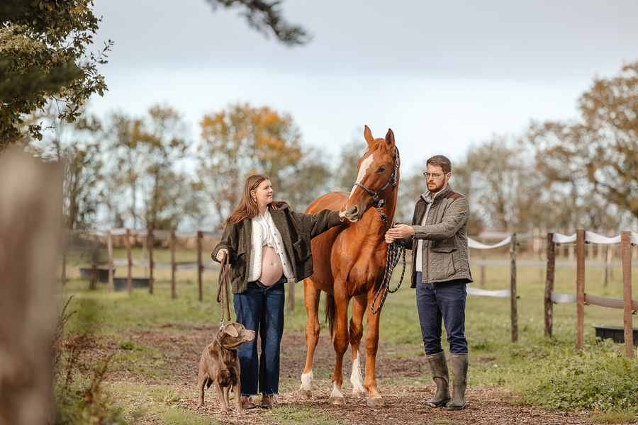 Femme enceinte, son compagnon et leur chien aux côtés de leur cheval dans un pré en Bretagne – séance photo équestre naturelle et lumineuse par Julie Séby Photographie