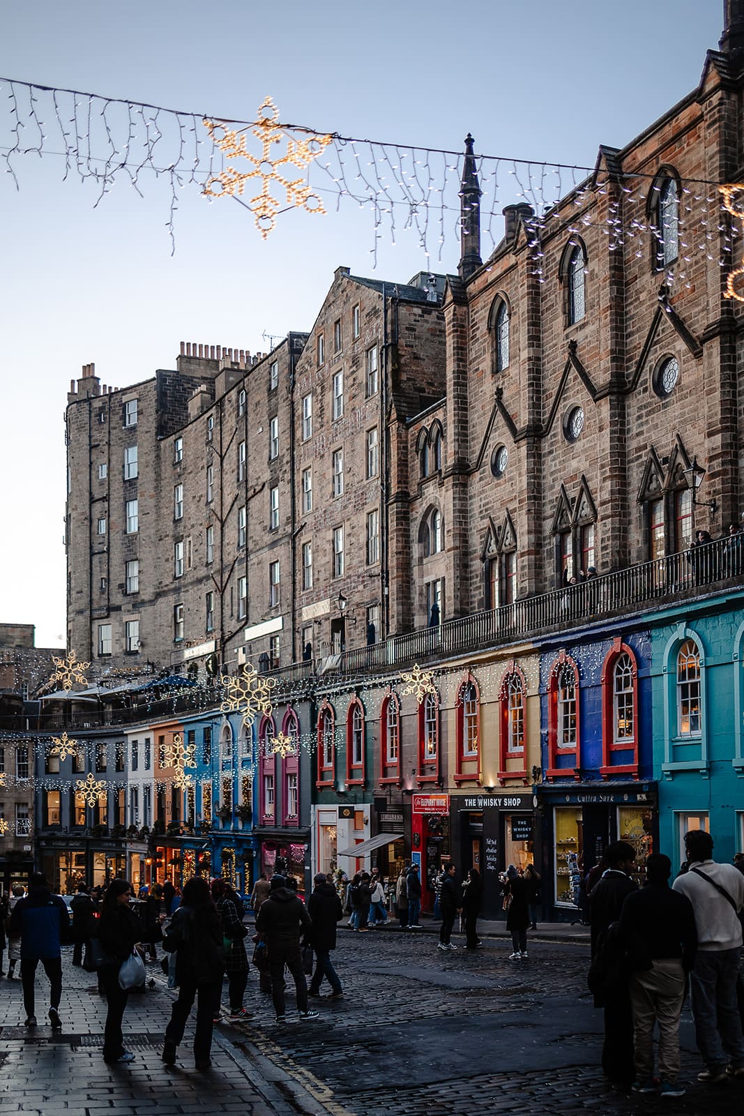 Rue courbe de Victoria Street avec ses façades colorées et ses décorations de Noël en forme de flocons lumineux suspendus au-dessus de la foule.