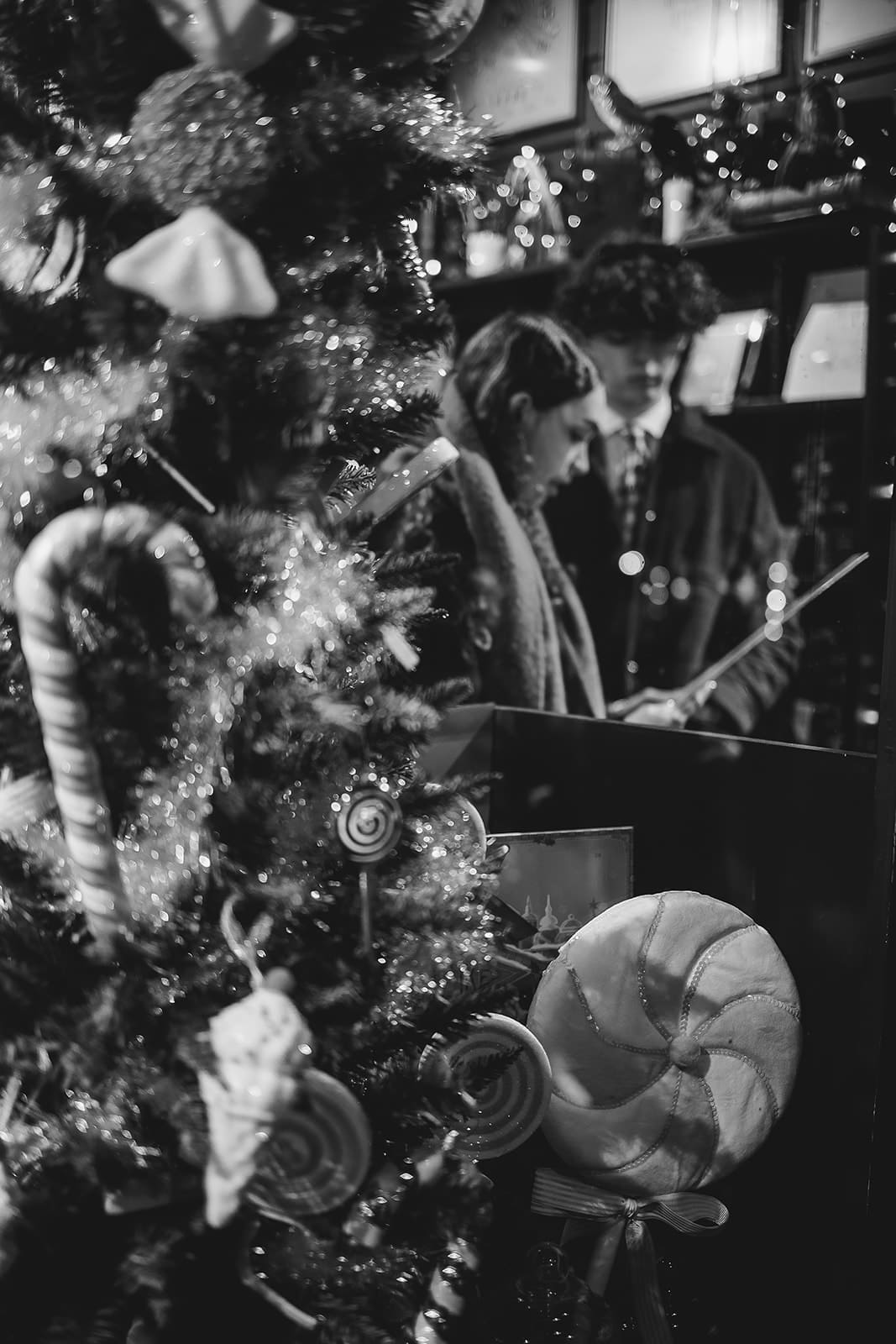 Photo en noir et blanc d'une vitrine de Noël avec un sapin décoré et des reflets de visiteurs devant la boutique MinaLima.