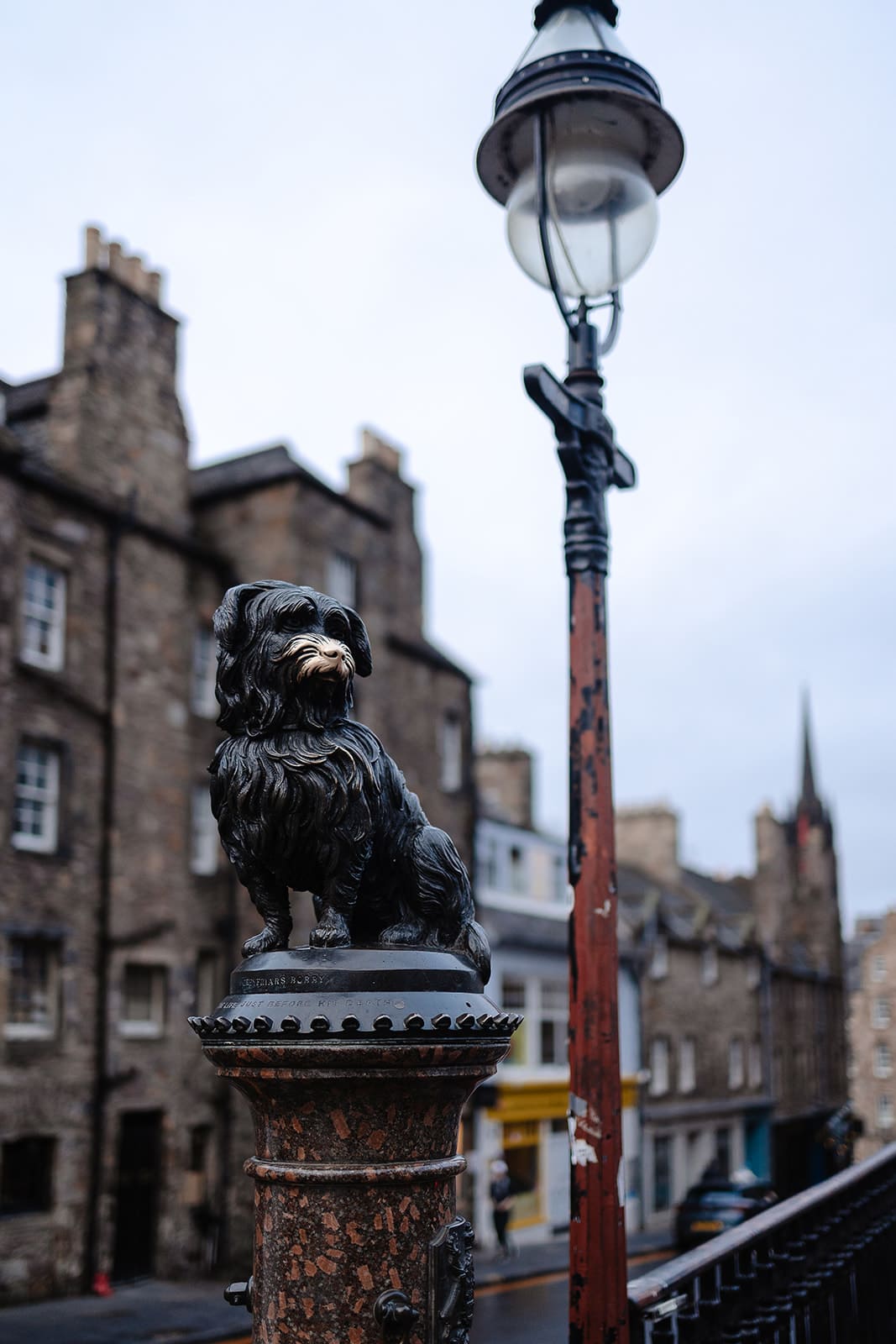 Statue du petit chien Greyfriars Bobby sur son socle, avec les bâtiments historiques de Candlemaker Row en arrière-plan.