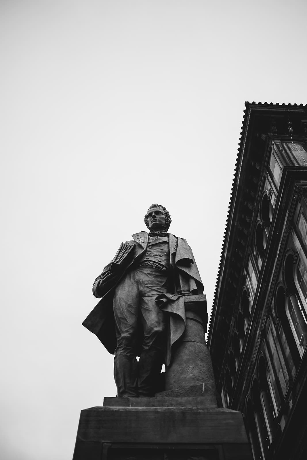 Statue en pied d'un homme illustre en habit d'époque, photographiée en contre-plongée noir et blanc à côté d'une façade classique.