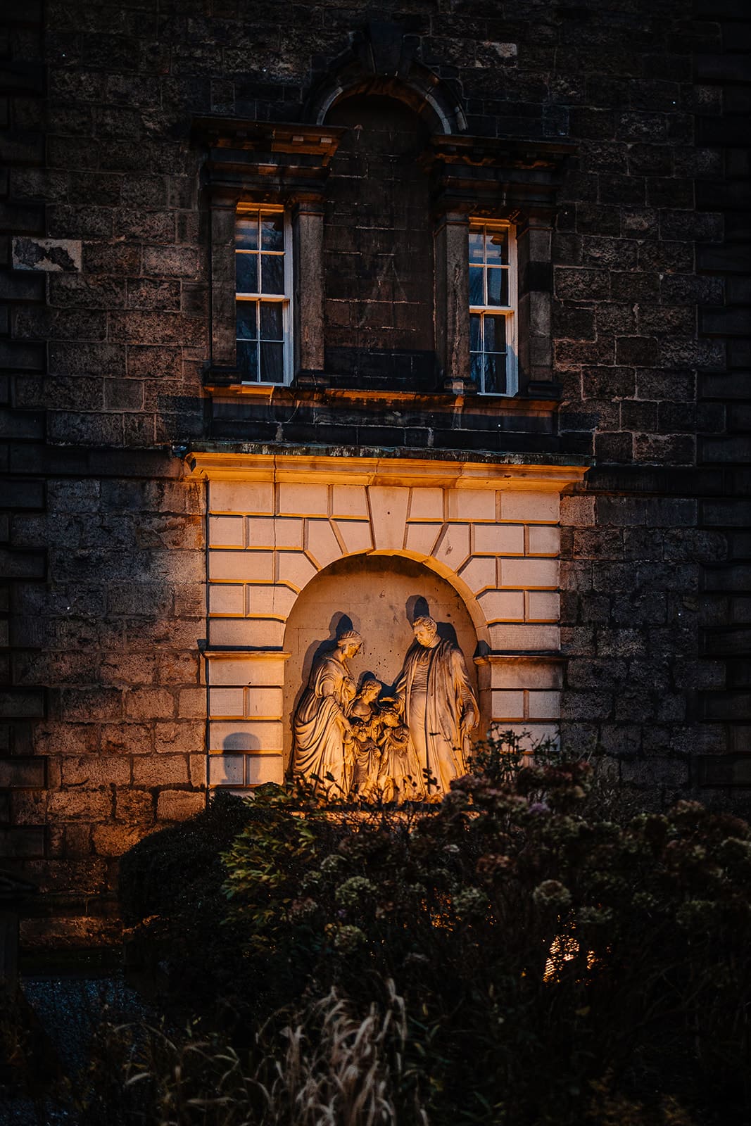Sculpture religieuse en bas-relief représentant trois personnages, incrustée dans un mur de briques en pierre et éclairée par une lumière chaude.