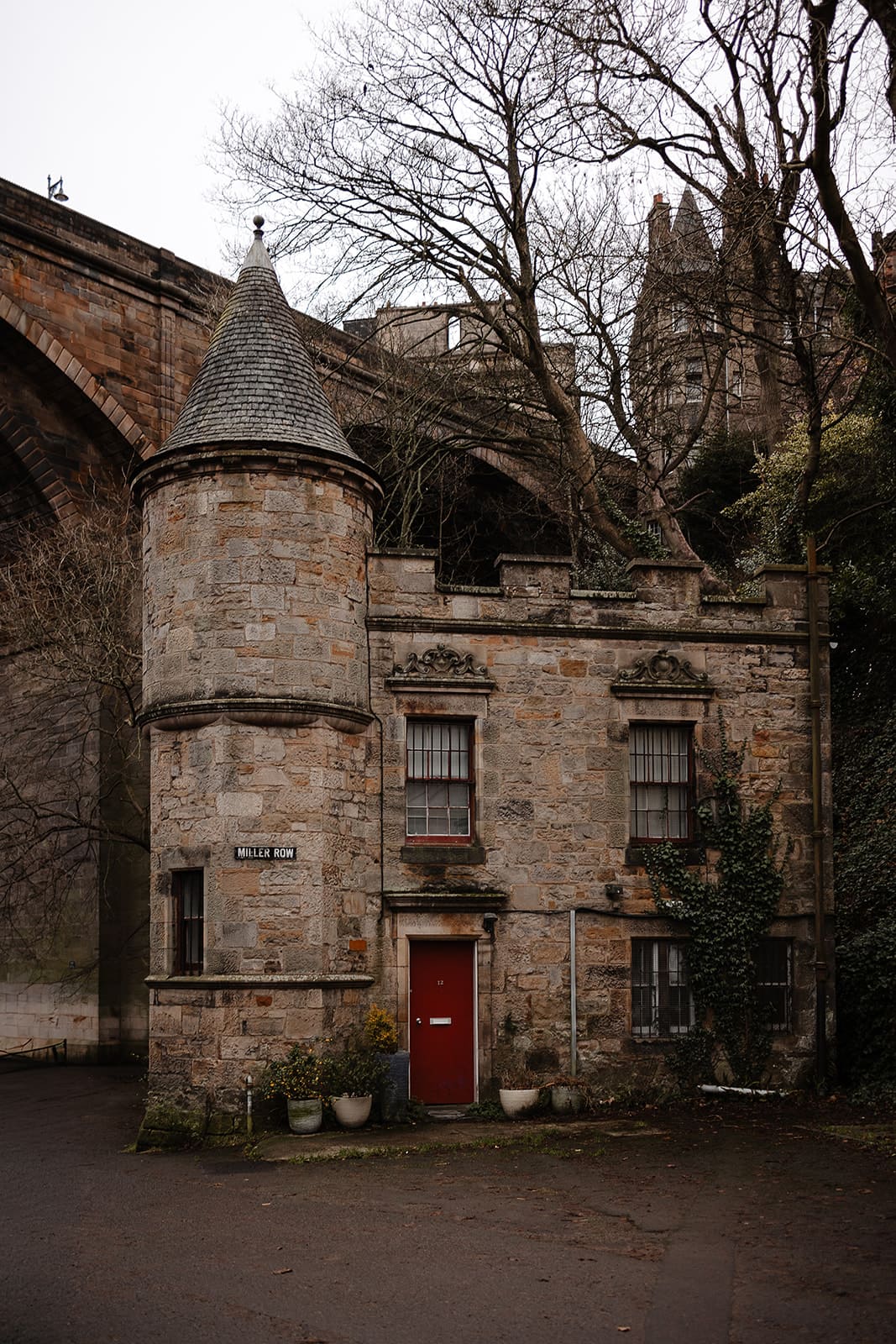 Petite maison ancienne en pierre avec une tour ronde à toit conique et une porte rouge, située sur Miller Row à Édimbourg sous un pont en arc.