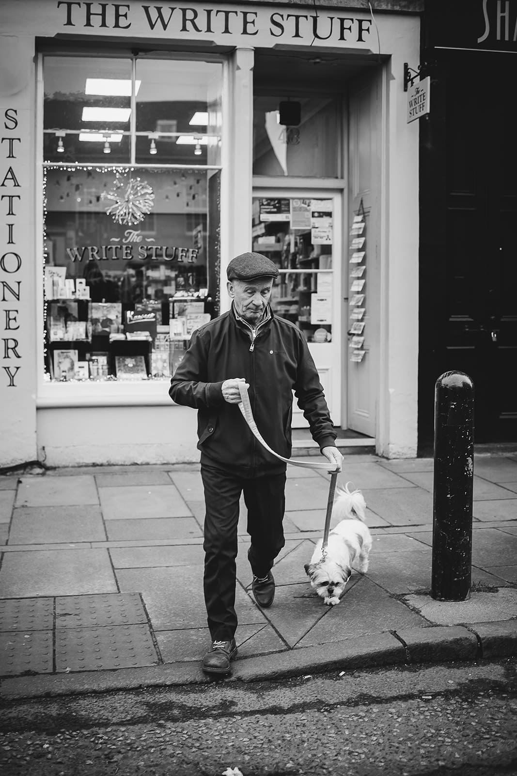 Photo de rue en noir et blanc d'un homme avec une casquette promenant son petit chien blanc devant une papeterie.