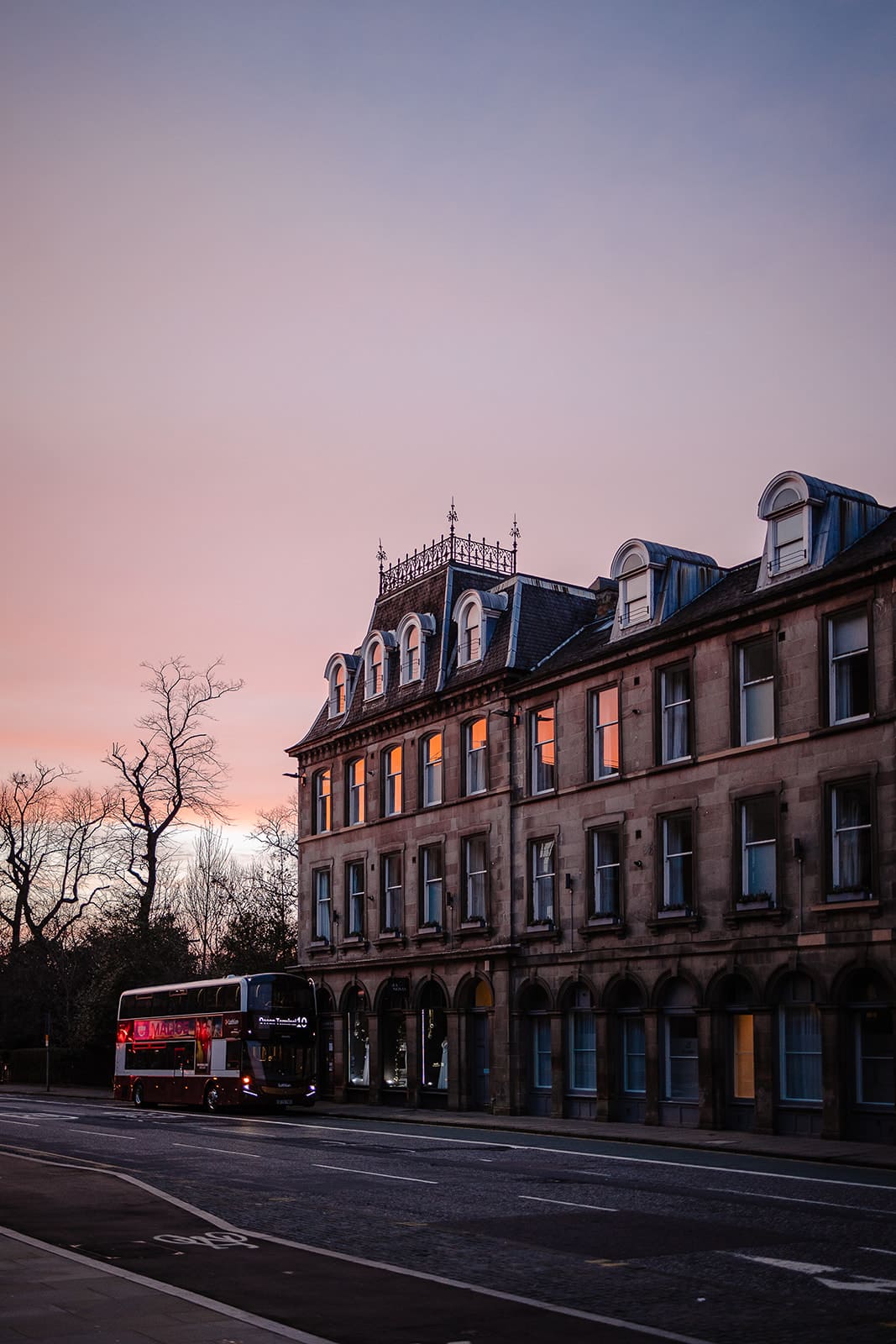 Immeuble écossais illuminé après le coucher du soleil avec un bus à impériale typique passant devant, créant une atmosphère urbaine nocturne.