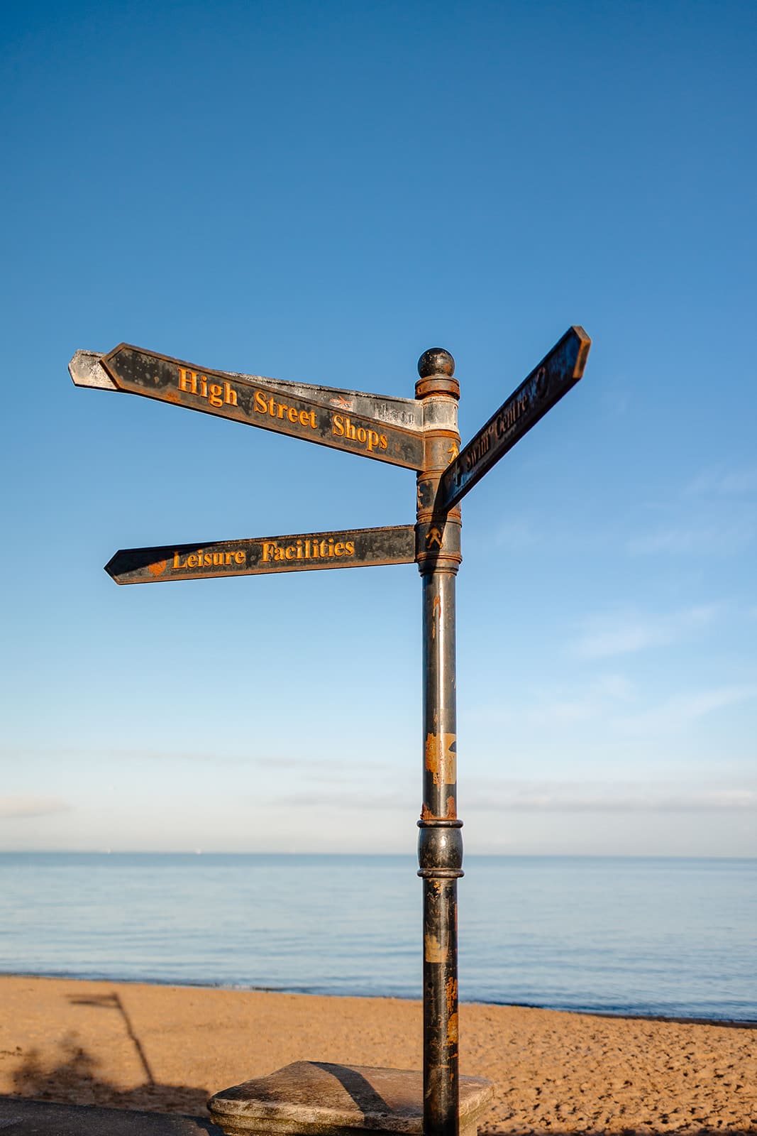 Panneau directionnel ancien en métal noir et doré sur la plage de Portobello à Édimbourg, indiquant "High Street Shops" et "Leisure Facilities" sous un ciel bleu pur.